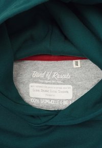 Grön bomullstyg med en mjuk textur, med en grå innertag som anger "100% Bomull (Ekologisk)" och märkesinformation.