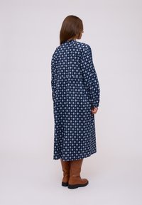 Donna in piedi di lato che indossa un abito midi a maniche lunghe blu navy con motivi a rombi bianchi e stivali marroni fino al ginocchio.