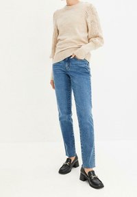 Person som bär en beige stickad tröja med mönstrade ärmar, blå raka jeans och svarta klackskor med gulddetaljer.