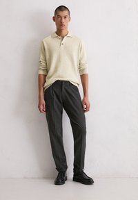 Lichtbeige gebreide polo sweater met geribbelde manchetten, gecombineerd met donkergrijze stijlvolle broek en zwarte veterschoenen. Gevoelige stofdetails zichtbaar.