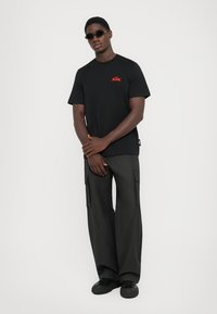 Sort T-shirt med korte ærmer og et rødt logo på brystet, parret med mørkegrå cargo-bukser med sidelommer.