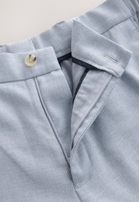 Helle blaue Stoffhosen mit strukturiertem Gewebe, ausgestattet mit einem Knopfverschluss und kontrastierendem Innenfutter. Sichtbare Nähte und glatte Kanten.