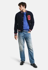 Giacca varsity di lana navy con polsini e colletto a coste blu e bianchi, con un'applicazione a forma di "G", abbinata a un maglione blue e jeans chiari.