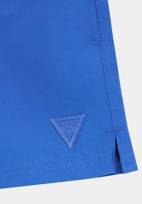 Pantaloni da bagno blu con una patch di plastica triangolare che riporta "GUESS BEACH" e un design di palma. Tessuto liscio con dettagli cuciti.