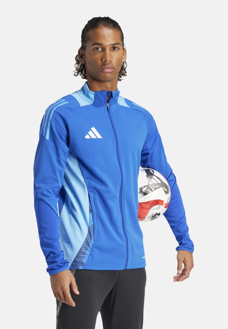 Blaue Sportjacke mit hellblauen Akzenten, durchgehendem Reißverschluss, stehender Kragen und gesticktem Logo. Das Modell hält einen Fußball.