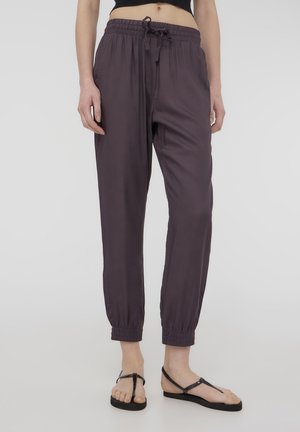 Persona che indossa pantaloni jogger viola scuro con coulisse e sandali neri piatti, in piedi davanti a uno sfondo semplice.