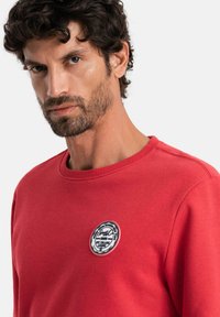 Sudadera roja hecha de una mezcla de algodón. Diseño de cuello redondo con un parche de logo circular en blanco y negro en el área del pecho izquierdo.