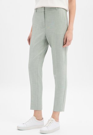 Personne portant un pantalon slim vert clair longueur cheville et des baskets blanches, debout devant un fond uni.