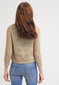 Beige stickad tröja med trekvartsärmar, öppet vävd design, ribbat nederkant och matchad med denimjeans. Bakifrån visad.