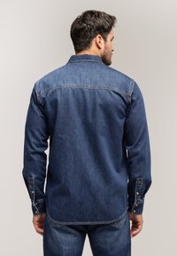 Denimowa koszula w ciemnoniebieskim kolorze z długimi rękawami, zapięciem na guziki i kołnierzykiem. Posiada ozdobne przeszycia oraz prosty dół.