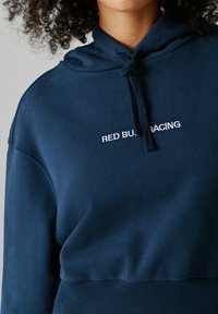 Osoba nosiace tmavomodrú mikinu s kapucňou, na ktorej je na hrudi vyšívaný nápis "RED BULL RACING" a kapucňa má štýlové šnúrky.