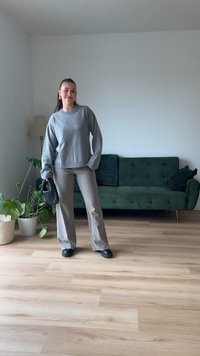 Grauer Pullover, beige weite Hosen und schwarze Schuhe. Innenraum mit grünem Sofa und Topfpflanze. Helle Holzböden und weiße Wände.