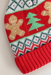 Red knitted winter hat featuring gingerbread men, a green Christmas tree, and a patterned cuff in white, green, and red. Soft texture.