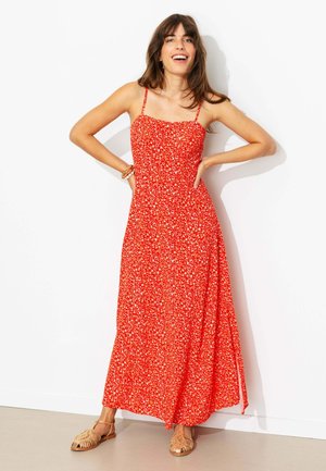 Femme aux longs cheveux bruns souriante, portant une robe longue rouge fleurie sans manches et des sandales plates tressées, debout les mains sur les hanches.