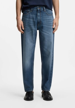 Mand iført blå straight-leg jeans, sorte sko og en mørk skjorte, stående mod en ensartet lys baggrund.