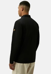 Zwarte langemouwen shirt van lichtgewicht stof, met een puntige kraag en een merkpatch op de linker mouw.