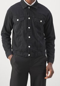 Zwarte spijkerjapon met een kraaghals, twee borstzakken, zilverkleurige drukknopen en een losse pasvorm, gestyled met geribbelde zwarte broek.