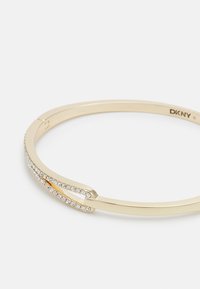 Pulsera bangle de tono dorado con un diseño elegante que presenta una fila de cristales transparentes a lo largo de un lado y un acabado suave y pulido.