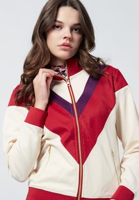 Giacca color-block in crema, rosso e viola. Presenta una chiusura con zip, colletto alto, polsini a costine e tasche laterali con dettagli in hardware dorato.