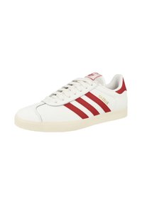 White leather sneakers with three red stripes on the side, a textured outsole, and a red heel accent. “Gazelle” logo in gold on the side.
