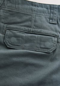 Gros plan d'une poche arrière en tissu gris avec un rabat et des coutures visibles sur un pantalon.