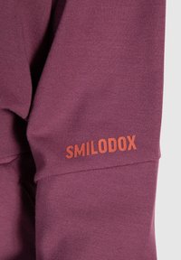 Felpa bordeaux in tessuto texturizzato, con il logo arancione "SMILODOX" visibile sulla manica sinistra in basso. Design semplice e aderente.