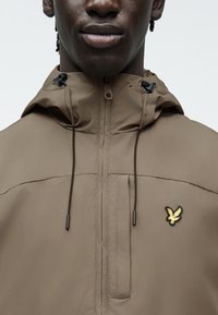 Κοντινό πλάνο ατόμου που φοράει καφέ μπουφάν με κουκούλα και φερμουάρ, με μικρό κεντητό κίτρινο λογότυπο πουλιού στο στήθος.