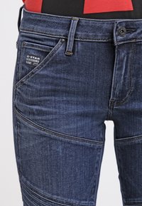 Jeans de denim azul oscuro con corte ajustado, que presentan múltiples costuras, un bolsillo de parche y una etiqueta negra en la cintura. Tejido texturizado.