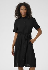 Robe noire à boutons avec manches courtes, ceinture à la taille et poches latérales. Le tissu semble lisse avec un design léger et fluide.