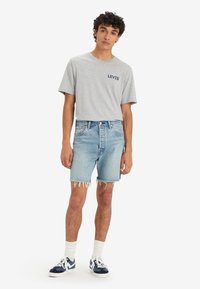 Camiseta de algodón gris con el logo en navy de "Levi's", combinada con pantalones cortos de mezclilla azul claro con dobladillos deshilachados. El modelo lleva zapatillas azules y calcetines blancos.