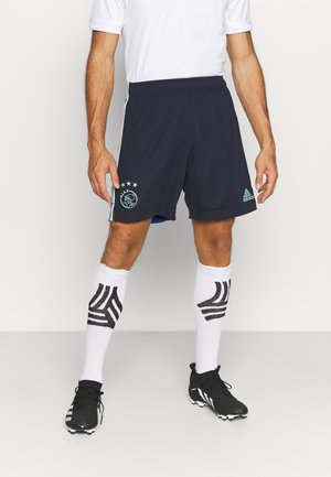 Homme portant un short de football Ajax bleu marine, des chaussettes blanches avec des rayures noires, des crampons noirs et un t-shirt blanc, debout sur un sol neutre.