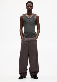 Canottiera grigia abbinata a jeans larghi di colore marrone scuro con cuciture visibili. Scarpe nere lucide completano l'outfit.