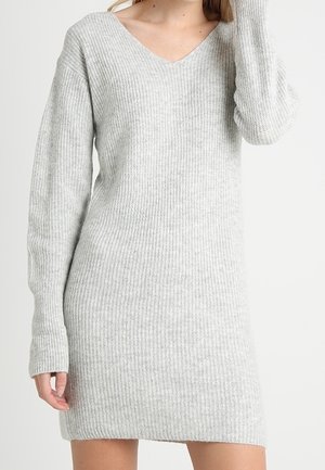 Vrouw draagt een lichtgrijze geribde gebreide sweaterjurk met lange mouwen en een V-hals, staand tegen een effen achtergrond.