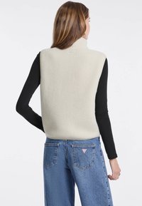 Cremfarbener Fleece-Westen mit hohem Kragen, weicher Textur und ohne Ärmel, getragen über einem schwarzen Langarmshirt mit blauen Jeans.