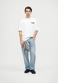 Joven de pie vistiendo una camiseta blanca de gran tamaño, jeans anchos de color azul claro con una bufanda en blanco y negro colgando del bolsillo, y zapatos negros.