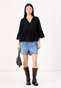 Black, textured top with a V-neck and button detail, paired with blue denim shorts and tall brown leather boots. Carrying a dark bag.