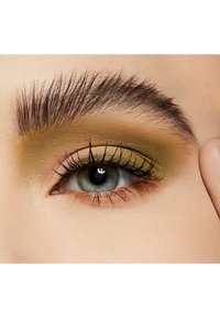 Close-up van een oog met olijfgroene en gouden oogschaduw, dikke wimpers en goed gedefinieerde wenkbrauwen tegen een lichte huidskleur achtergrond.
