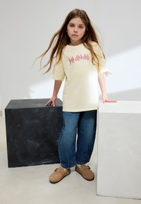 Cremefarbenes oversized T-Shirt mit rotem "Def Leppard"-Logo, kombiniert mit blauen Jeans aus Denim und braunen Clogs, stehend zwischen hellen und dunklen Blöcken.