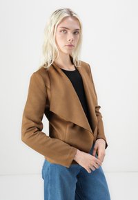 Brauner, weicher Stoffblazer mit offenem vorne und langen Ärmeln, kombiniert mit einem schwarzen Oberteil und blauen Jeans. Glatte Textur, strukturiertes Design.