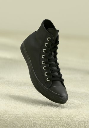 Czarny wysoki sneaker z czarnymi sznurowadłami i metalowymi oczkami, ustawiony pod kątem na miękkiej, kremowej, teksturowanej powierzchni.