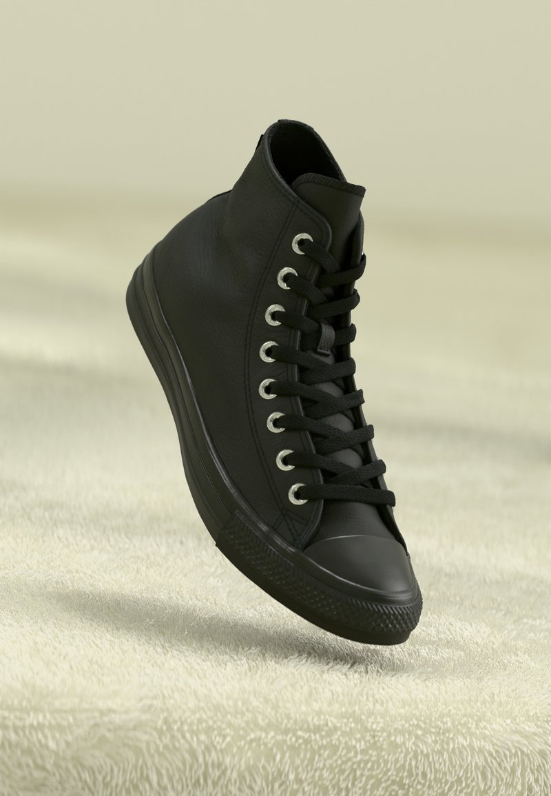 Basket montante noire avec des lacets noirs et des œillets métalliques, inclinée vers le haut sur une surface texturée douce de couleur crème.