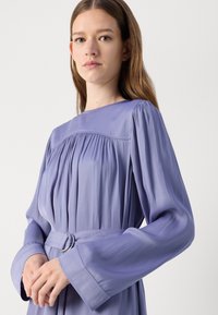 Abito a maniche lunghe di lavanda con dettagli plissettati, scollatura alta e vita con cintura e fibbia rotonda in argento. Tessuto dalla consistenza liscia.