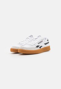 White leather sneakers with black accents, suede toe, and gum rubber outsole. Features lace-up closure and Reebok branding on side.