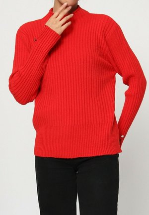 Pull en maille côtelée rouge avec col montant, manches longues et coupe légèrement ample. Présente un petit accent décoratif sur l'épaule gauche.