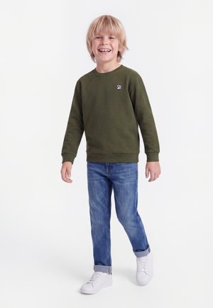 Smilende blond dreng går, iført en olivengrøn sweatshirt, opsmøgede blå jeans og hvide sneakers på en hvid baggrund.