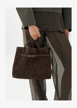 Person holding a dark brown suede handbag with double handles and a thin belt detail, wearing sheer brown pants and a textured long-sleeve top.