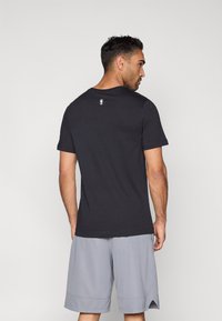 Μαύρο βαμβακερό t-shirt με κοντά μανίκια, στρογγυλή λαιμόκοψη και μικρή λεπτομέρεια με λογότυπο στο πάνω μέρος της πλάτης. Φοριέται με γκρι σορτς.