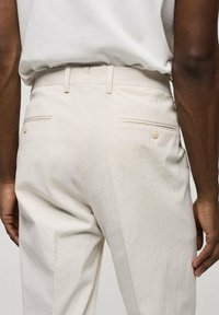 Světlé béžové kalhoty vyrobené z texturované tkaniny, s dvěma zadními kapsami na knoflíky a pasem s poutky na opasek.