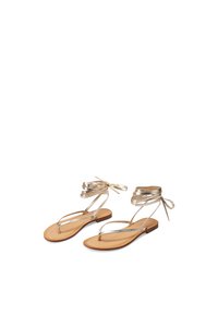 Sandali piatti dorati con laccetti alla caviglia, caratterizzati da un design minimalista, sottili cinturini e una suola in tessuto liscio color beige.
