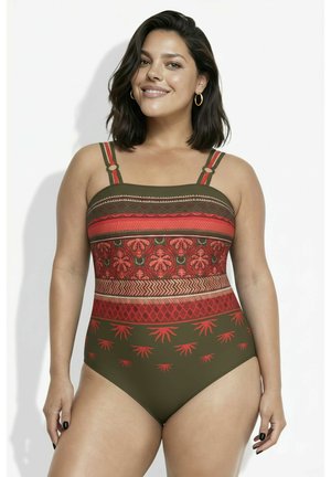 Uśmiechnięta kobieta o ciemnych włosach ubrana w oliwkowozielony jednoczęściowy strój kąpielowy z czerwonymi, pomarańczowymi i brązowymi poziomymi paskami.